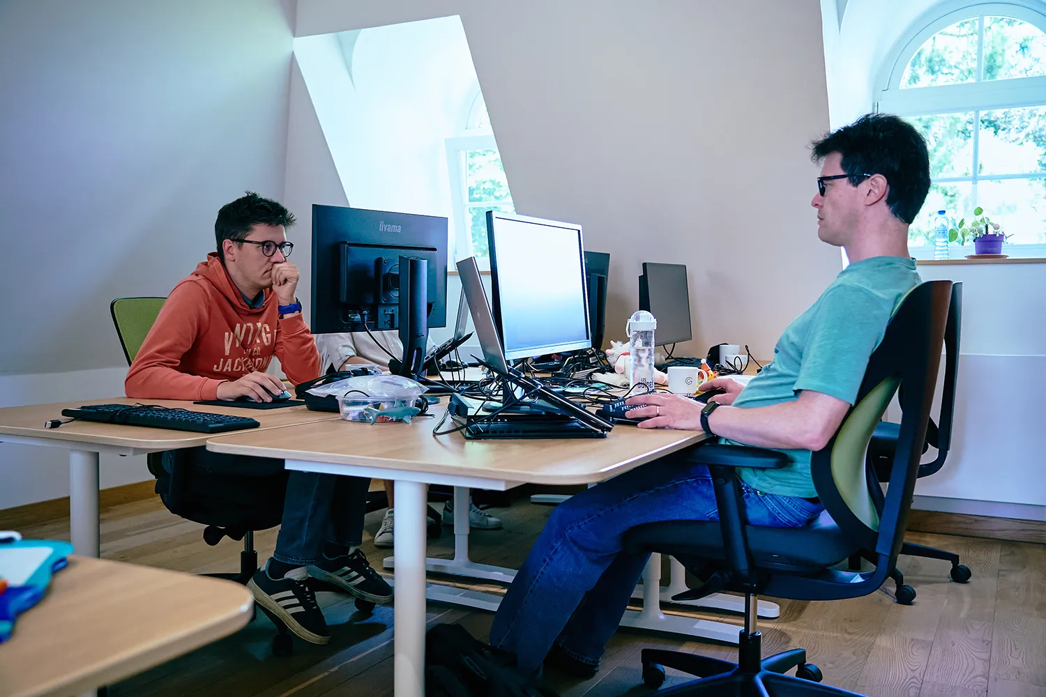 Deux hommes travaillent sur des ordinateurs dans un bureau lumineux, avec des écrans et divers accessoires sur les bureaux en bois.